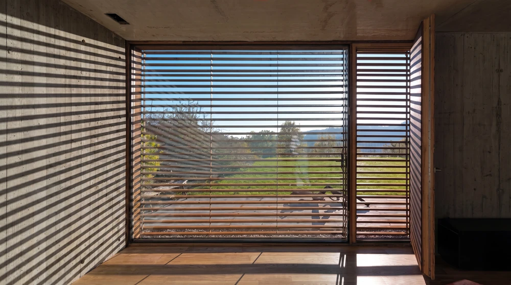 Vue intérieure d'une baie vitrée coulissante à grande surface, équipée de lames de brise-soleil orientables en bois foncé, montage horizontal