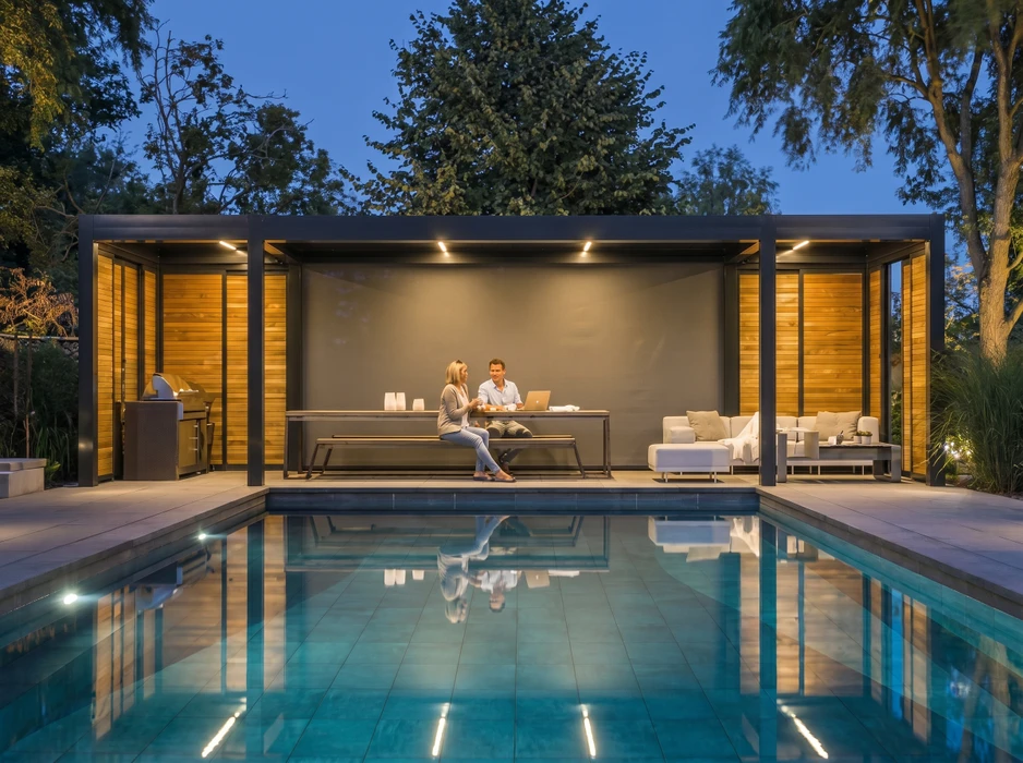Pool house moderne et contemporain construit en bois ou matériaux composites, directement adjacent à une piscine rectangulaire à liner. La structure est partiellement couverte avec terrasse ouverte en bois composite ou bois traité. À l'intérieur visible par les baies vitrées : espace de détente avec mobilier moderne. Autour : terrasse en bois qui relie le pool house à la piscine, végétation paysagère discrète. Ambiance lumineuse de jour ensoleillé. Photo prise en perspective montrant l'harmonie entre la structure, la piscine et l'environnement paysager.