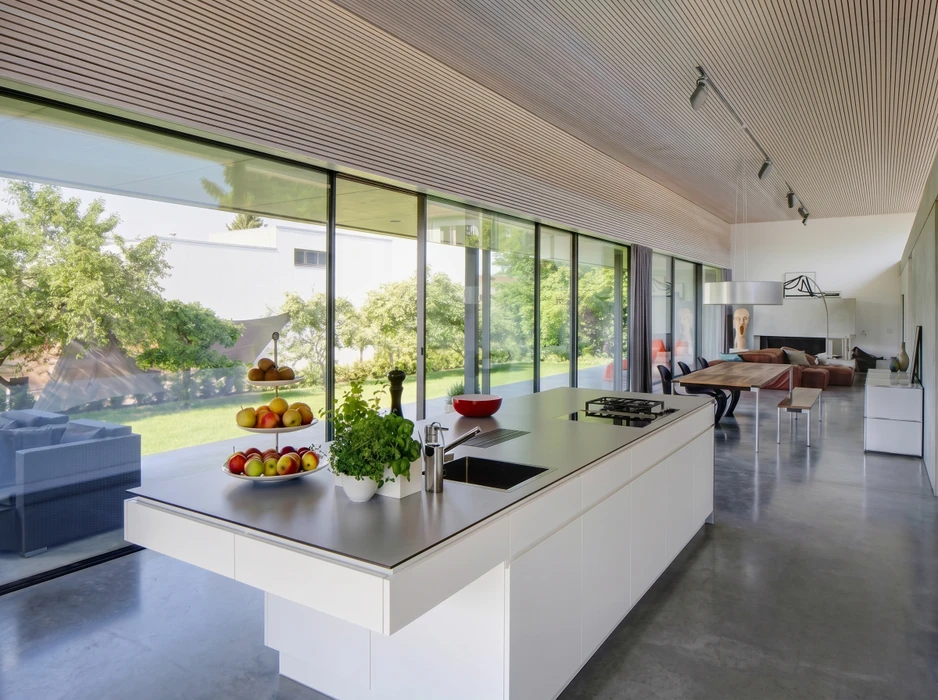 Photographie réaliste d'une véranda moderne construite en menuiserie aluminium et verre, intégrée à une maison résidentielle. La véranda est bien éclairée par la lumière naturelle, mettant en valeur la structure élégante des châssis et la transparence des vitres. L'espace intérieur accueille du mobilier de détente (chaise longue ou assises) offrant une vue dégagée sur le jardin extérieur. La pose et la finition professionnelle des menuiseries doivent être clairement visibles.