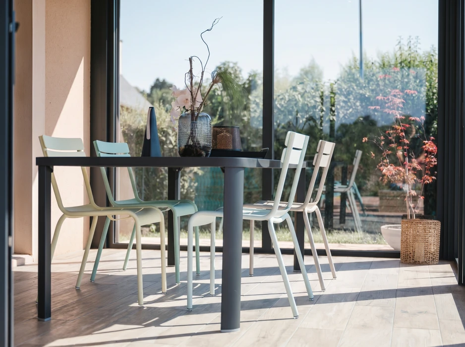 Photographie réaliste d'une véranda moderne construite en menuiserie aluminium et verre, intégrée à une maison résidentielle. La véranda est bien éclairée par la lumière naturelle, mettant en valeur la structure élégante des châssis et la transparence des vitres. L'espace intérieur accueille du mobilier de détente (chaise longue ou assises) offrant une vue dégagée sur le jardin extérieur. La pose et la finition professionnelle des menuiseries doivent être clairement visibles.