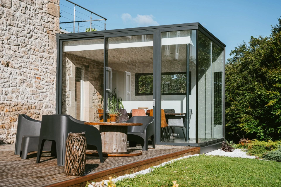 Véranda moderne avec structure aluminium et panneaux vitrés, mobilier en résine tressée noire, vue sur jardin paysager avec pelouse, massifs fleuris de tulipes roses, muret en pierre naturelle et clôture bois composite