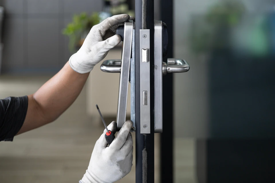 Technicien en salopette rouge et t-shirt blanc installe une serrure sur porte d'entrée en bois foncé dans hall d'immeuble moderne
