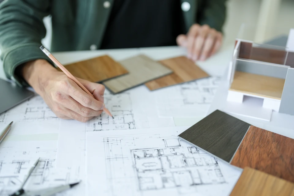 Un artisan menuisier ou professionnel de Génie Créations en conversation avec un client dans le showroom, présentant des plans ou des échantillons de fermetures (fenêtres, volets, portes) et de protections solaires (stores, pergolas). La scène montre une relation de confiance et de suivi personnalisé, avec le client examinant attentivement les matériaux aux côtés du professionnel qui explique les détails du projet.
